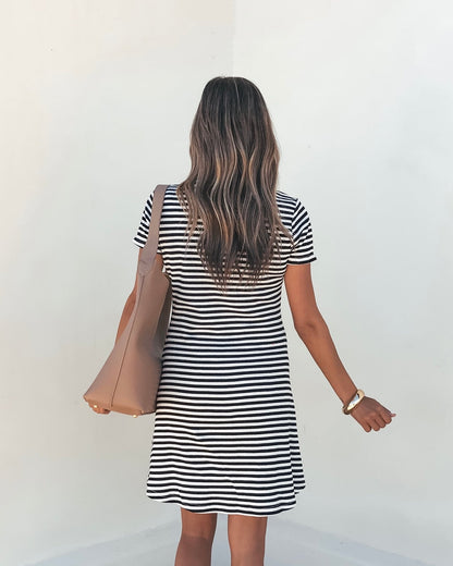 Woman with long wavy hair wears the Lucy Black and White Striped Mini Dress-FINAL SALE, holding a tan bag by a white wall.
