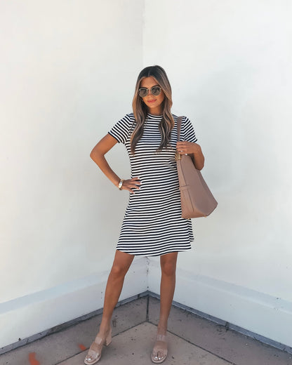 Woman in the Lucy Black and White Striped Mini Dress stands against a white wall, accessorized with sandals, sunglasses, and a tan handbag.