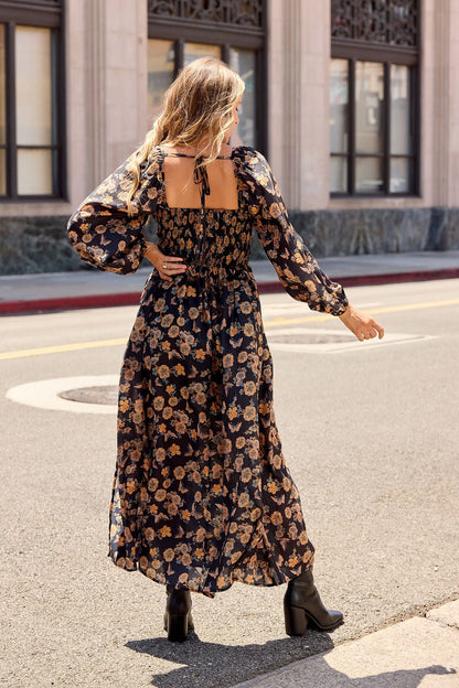 A woman in a Maple Floral Maxi Dress - Black and black boots walks down a city street, back to camera.