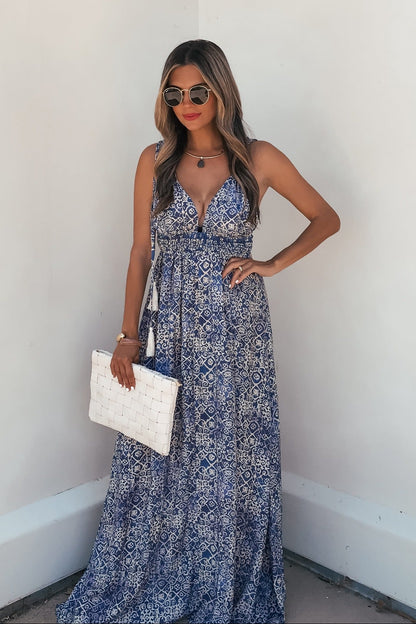 Woman in the Maribel Blue Print Maxi Dress - FINAL SALE, sunglasses, and white purse stands by a white wall.