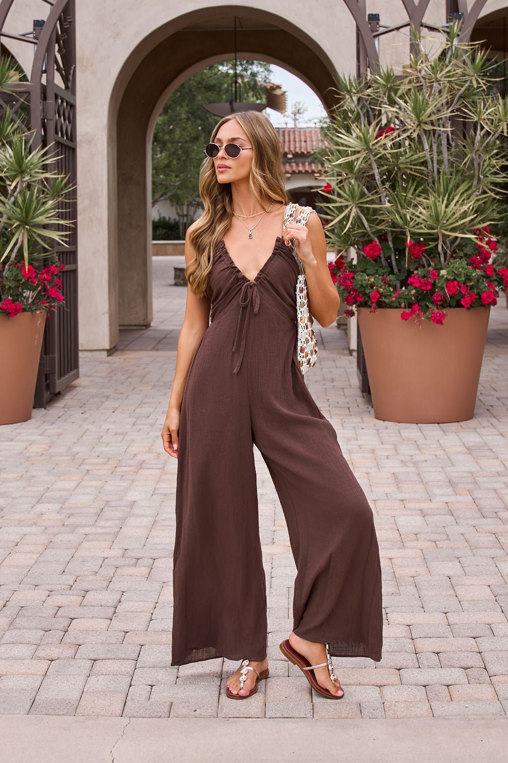 Woman in Marisol Brown Tie‑Front Wide Leg Jumpsuit stands outdoors by potted plants and arches, wearing sandals and sunglasses.