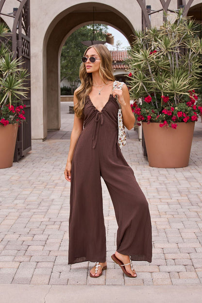 Woman in Marisol Brown Tie‑Front Wide Leg Jumpsuit stands outdoors by potted plants and arches, wearing sandals and sunglasses.