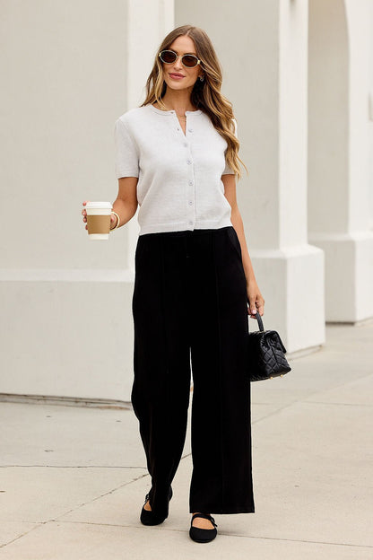 Woman in sunglasses, Maura Grey Short Sleeve Sweater Cardigan, and black wide-leg pants walks outside with coffee and a small black purse.
