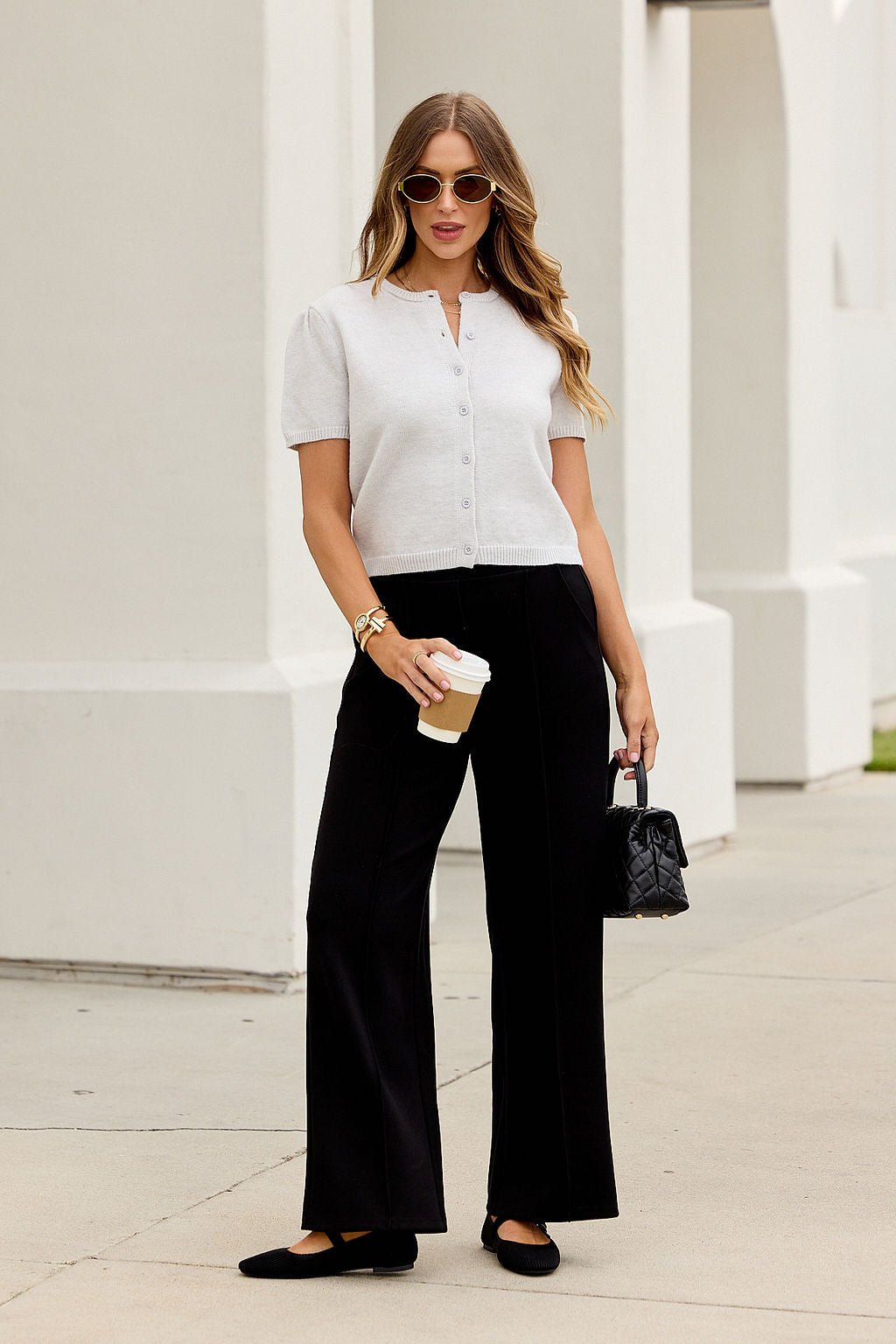 Woman in sunglasses with wavy hair wears the Maura Grey Short Sleeve Sweater Cardigan, black pants, flats, and holds coffee and a small purse.