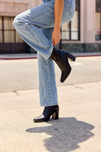 Someone in blue jeans adjusts their black MIA Sidonie Leather Booties while standing on a sunny sidewalk.