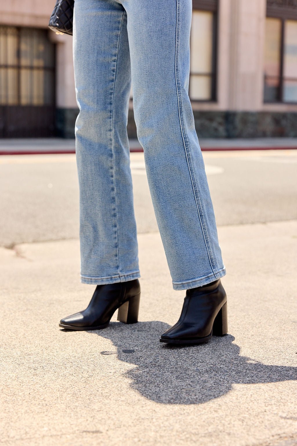 Person in light blue jeans and MIA Sidonie Leather Booties - Black stands on a city sidewalk, casting a shadow in the afternoon light.