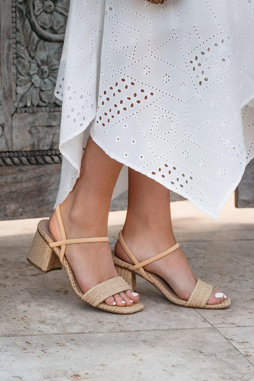 Woman in a white eyelet dress wears MIA Zissy Natural Strappy Heels-FINAL SALE, standing stylishly on stone flooring.
