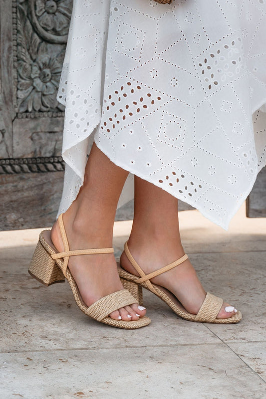 Woman in a white eyelet dress wears MIA Zissy Natural Strappy Heels-FINAL SALE, standing stylishly on stone flooring.