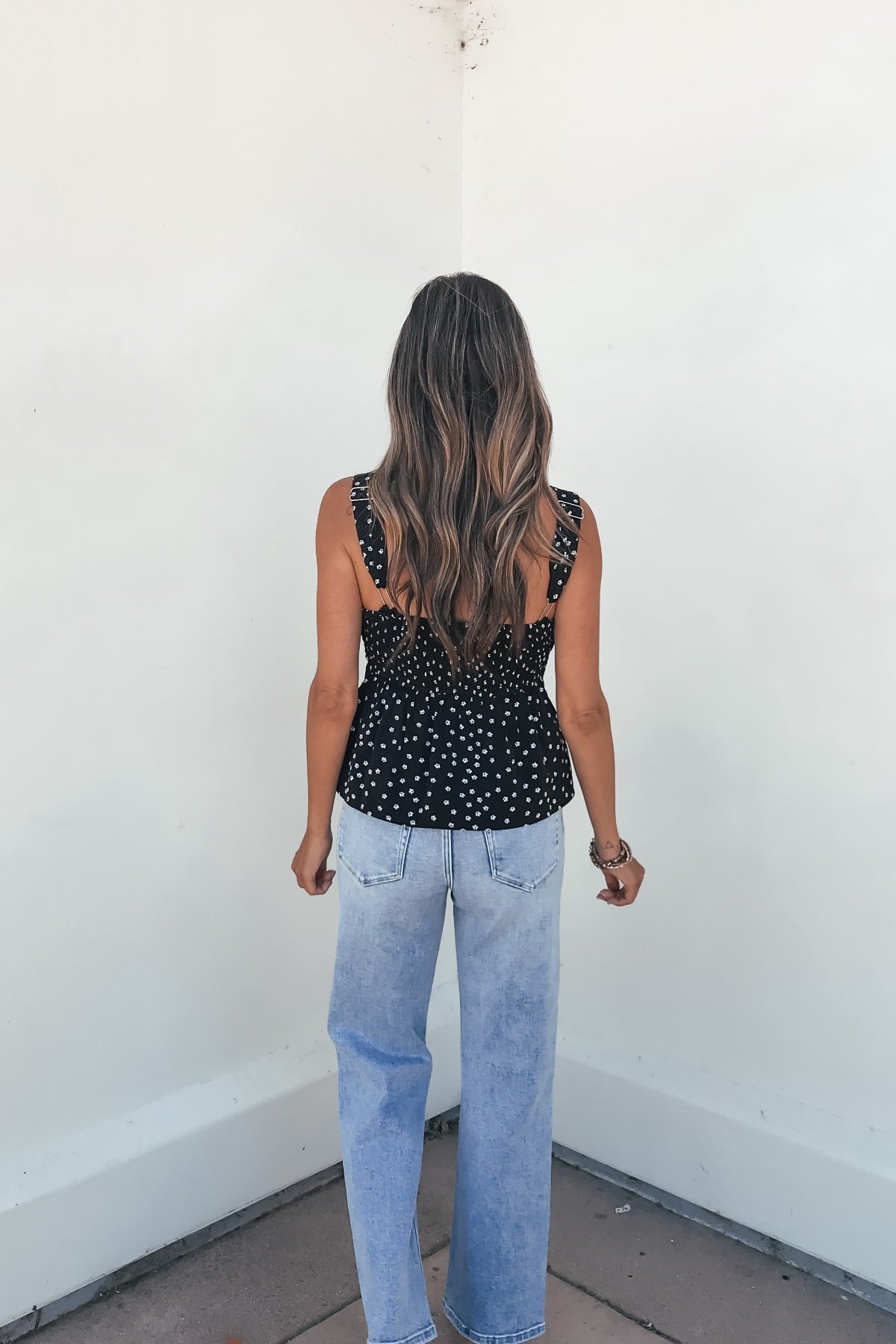 A woman with long wavy hair wears the Midnight Lace Print Tank Top - Black, facing a white corner wall.