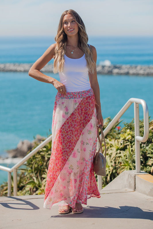 Woman in a white tank and Mixed Print Smocked Maxi Skirt - Pink stands outdoors by the ocean, smiling on a sunny day.