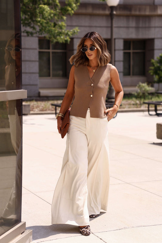 Woman in the Mocha Button Up Sweater Vest-FINAL SALE and white wide-leg pants walks outdoors with sunglasses and a clutch.