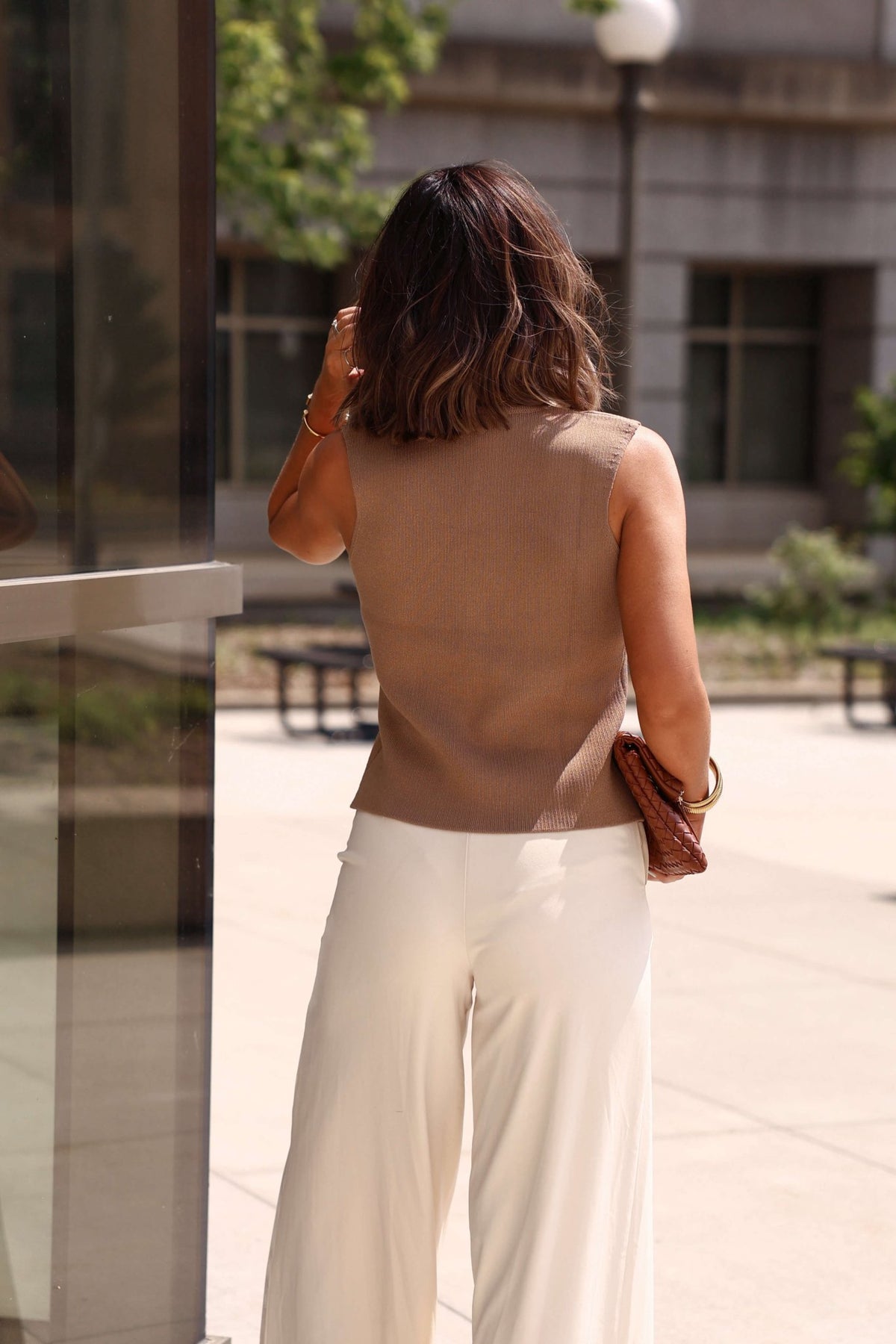 Woman with shoulder-length hair in the Mocha Button Up Sweater Vest-FINAL SALE and white pants stands outdoors, holding a brown clutch.