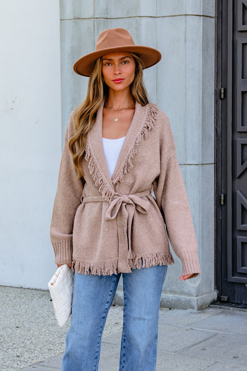 Woman in a tan hat, Mocha Fringe Tie Cardigan, white top, and jeans stands outdoors holding a chic white clutch.