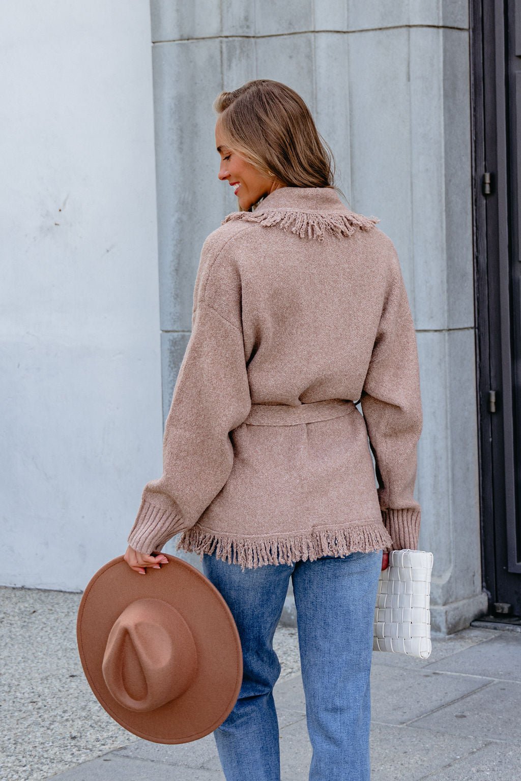 Woman in a Mocha Fringe Tie Cardigan and blue jeans, holding a brown hat and white clutch, stands outside by a building.