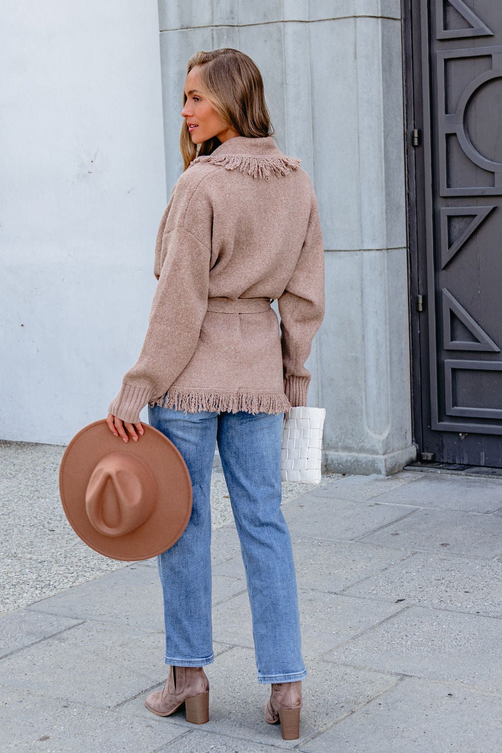 Woman in a Mocha Fringe Tie Cardigan and blue jeans holds a brown hat and white purse on a city sidewalk, facing away.