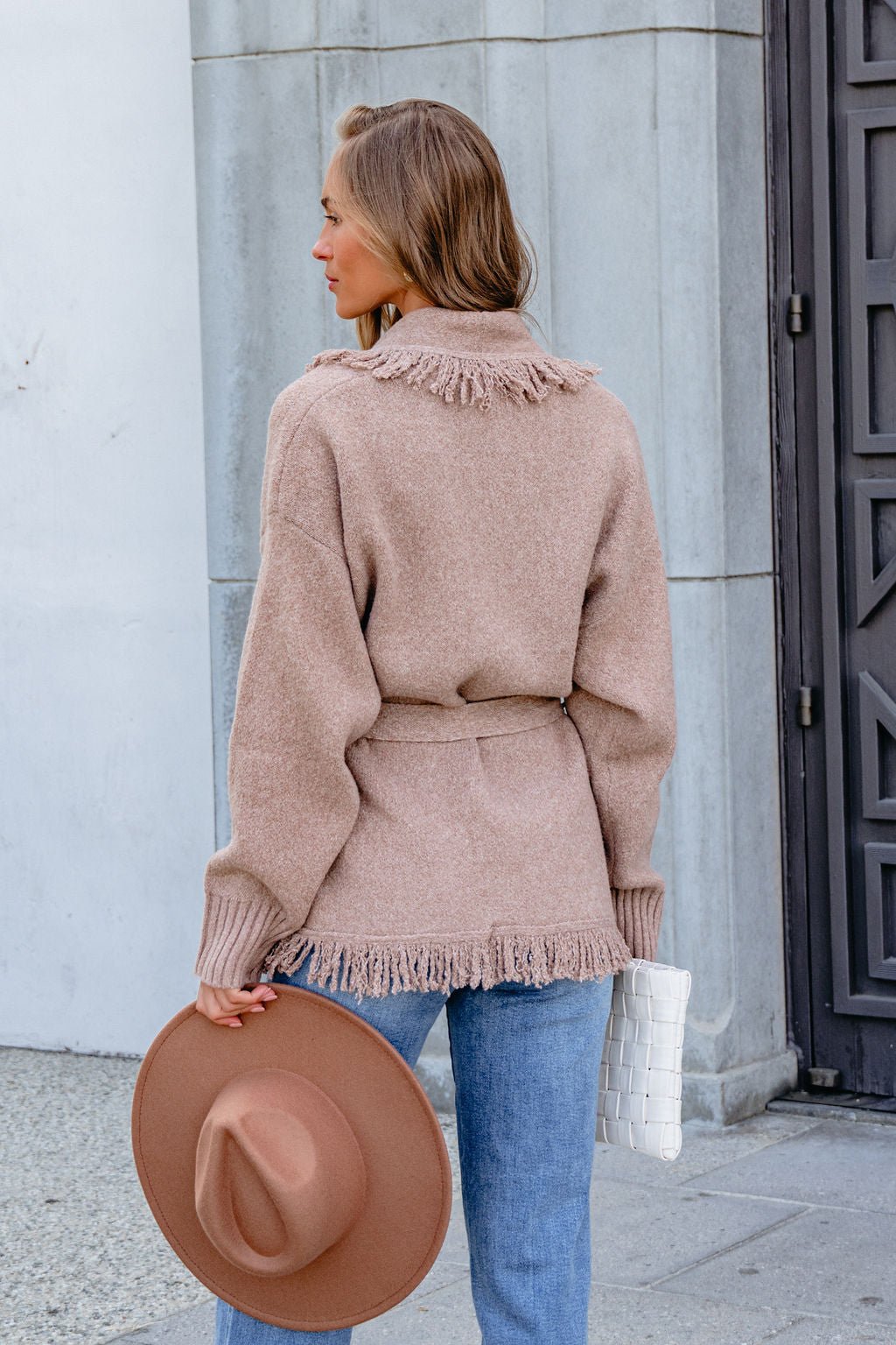 Woman in a Mocha Fringe Tie Cardigan holds a brown hat and white clutch, facing away outdoors.