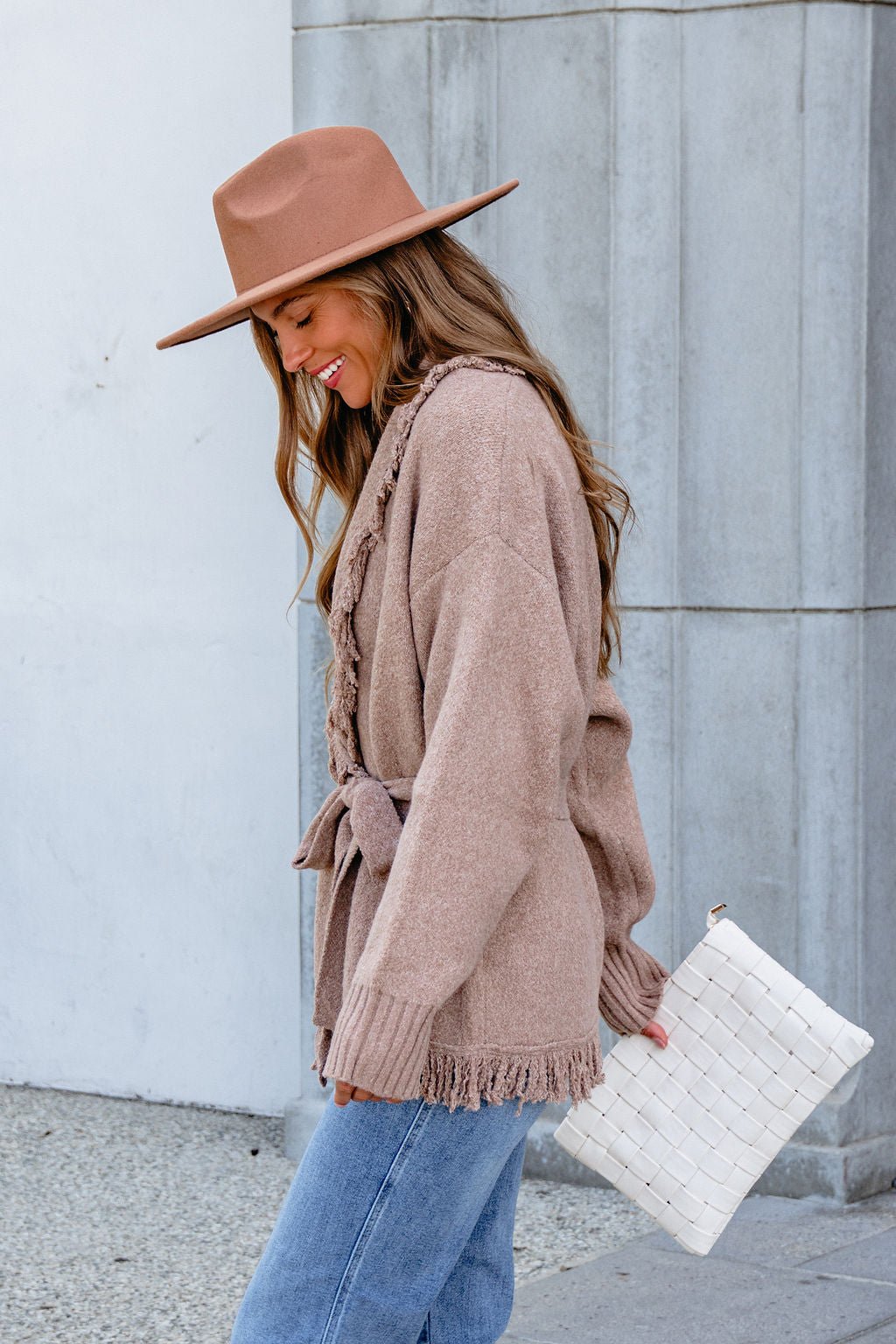 Smiling woman in a brown hat, jeans, and Mocha Fringe Tie Cardigan holds a white woven clutch while looking down.