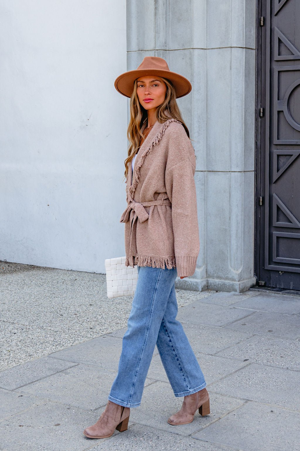Woman in a tan hat, Mocha Fringe Tie Cardigan, blue jeans, and ankle boots walking by a gray building.