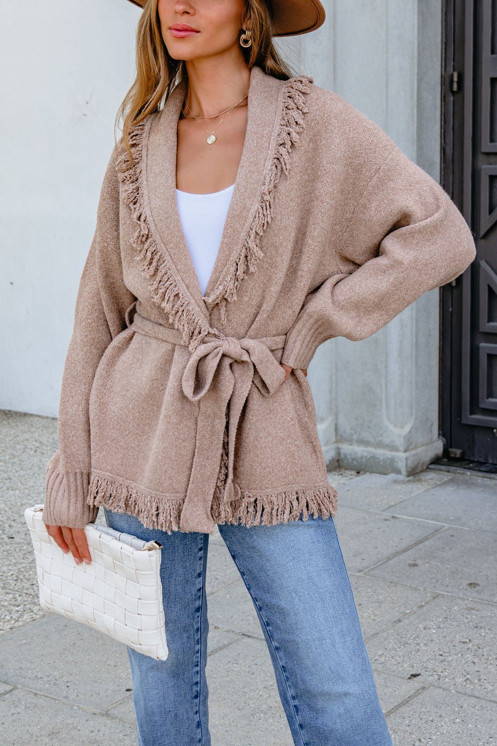 Woman in a Mocha Fringe Tie Cardigan, blue jeans, and a hat holds a white woven clutch while standing outdoors.