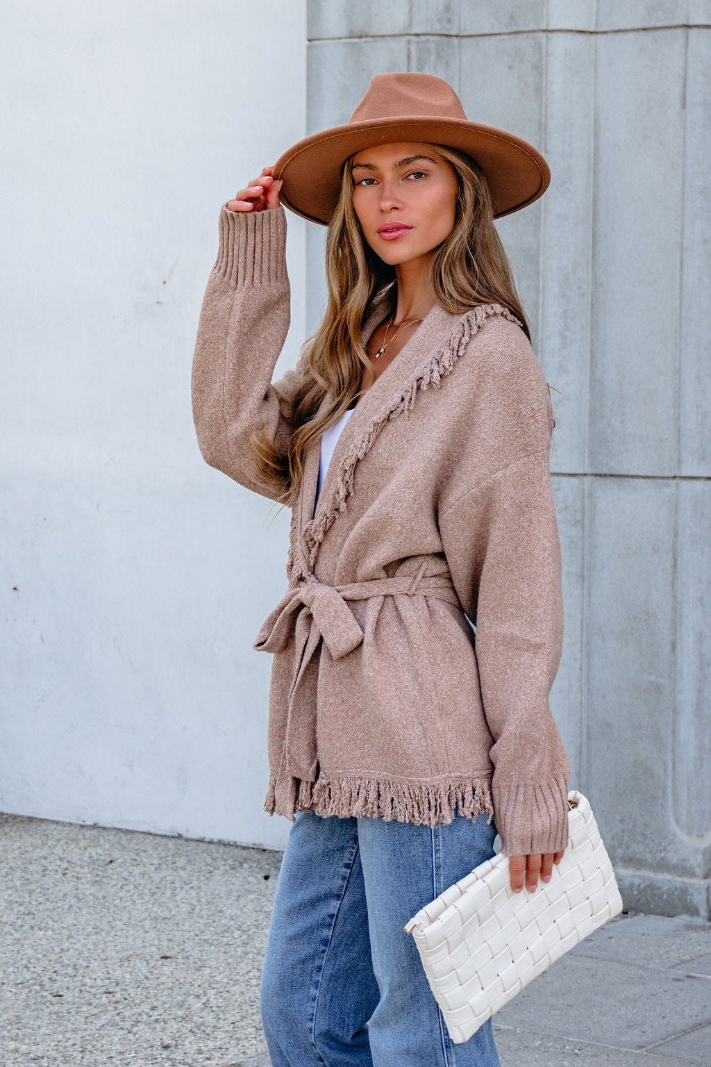 Woman in a brown hat, Mocha Fringe Tie Cardigan, and jeans holds a white clutch near a gray and white wall.