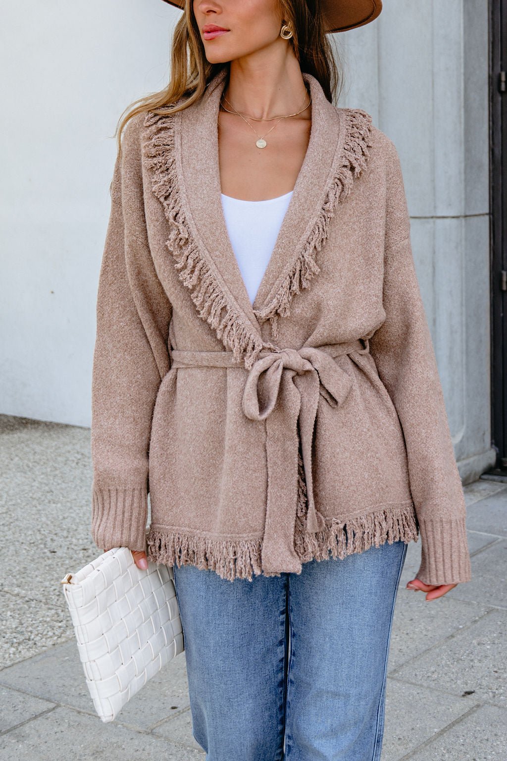Woman wearing a Mocha Fringe Tie Cardigan, blue jeans, and a white clutch, standing outdoors.