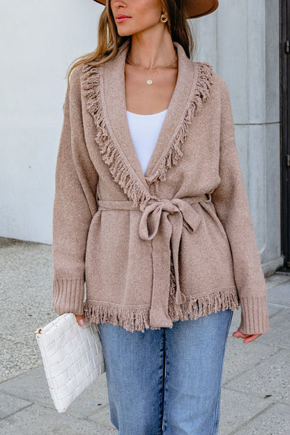 Woman wearing a Mocha Fringe Tie Cardigan, blue jeans, and a white clutch, standing outdoors.