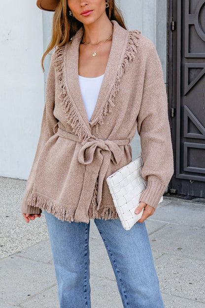 Woman in a Mocha Fringe Tie Cardigan, white top, blue jeans, and white clutch stands outdoors with effortless chic style.