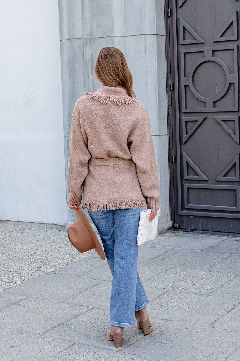 Person in a Mocha Fringe Tie Cardigan, blue jeans, and heels stands outdoors holding a brown hat and white clutch, facing away.