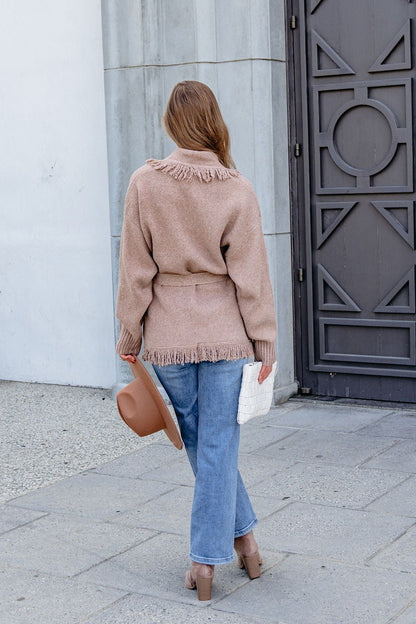 Person in a Mocha Fringe Tie Cardigan, blue jeans, and heels stands outdoors holding a brown hat and white clutch, facing away.