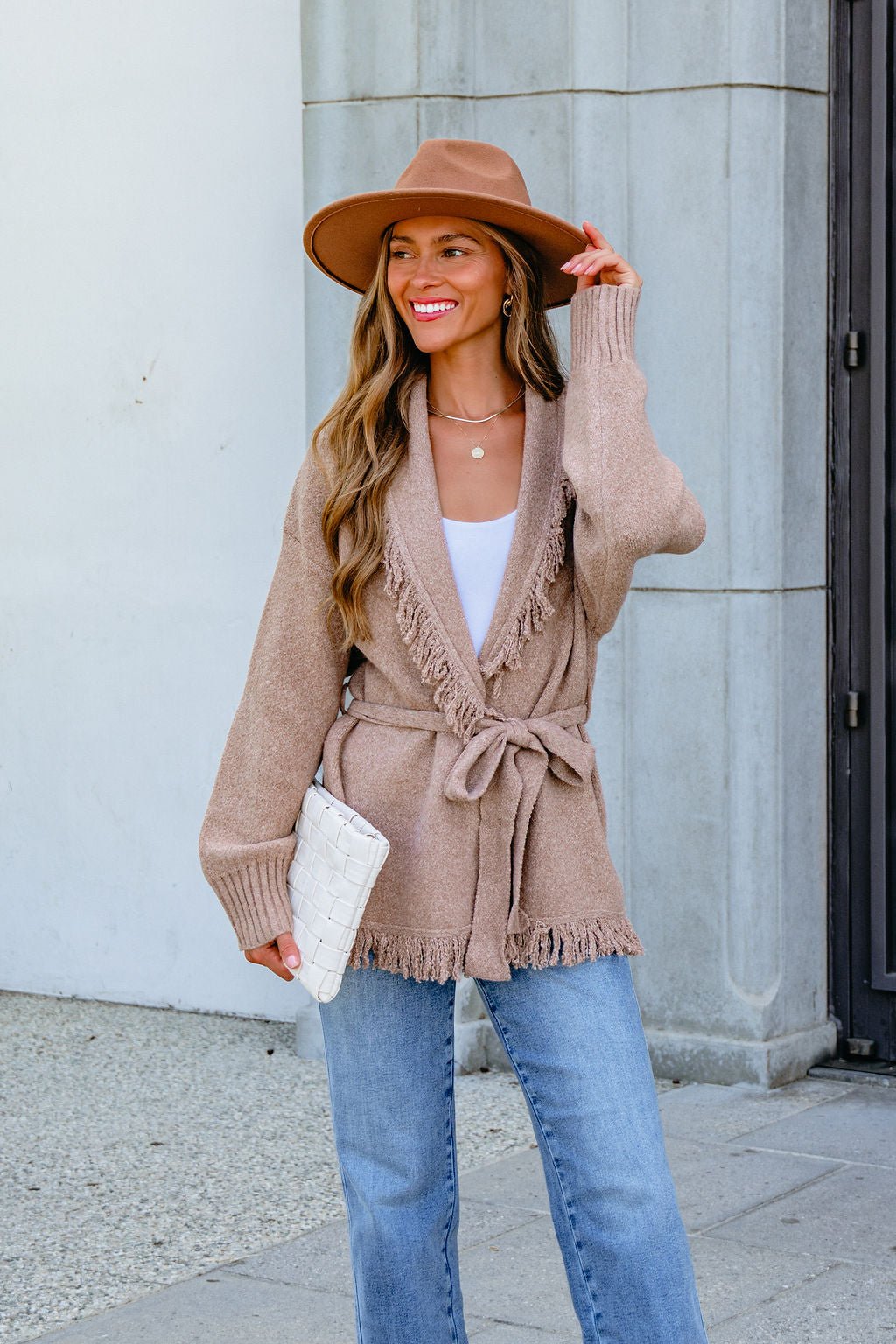 Woman in a tan hat, Mocha Fringe Tie Cardigan, white top, blue jeans, holding a white clutch, smiling outdoors.
