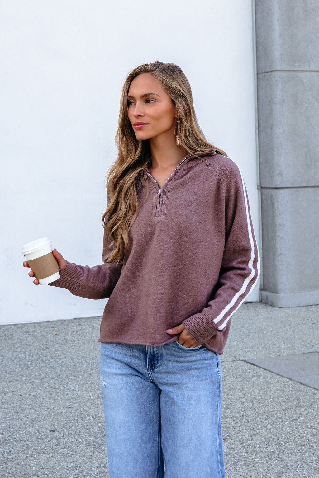 Woman with long hair wears a Mocha Half Zip Collared Pullover and jeans, holding a coffee cup against a white and gray background.