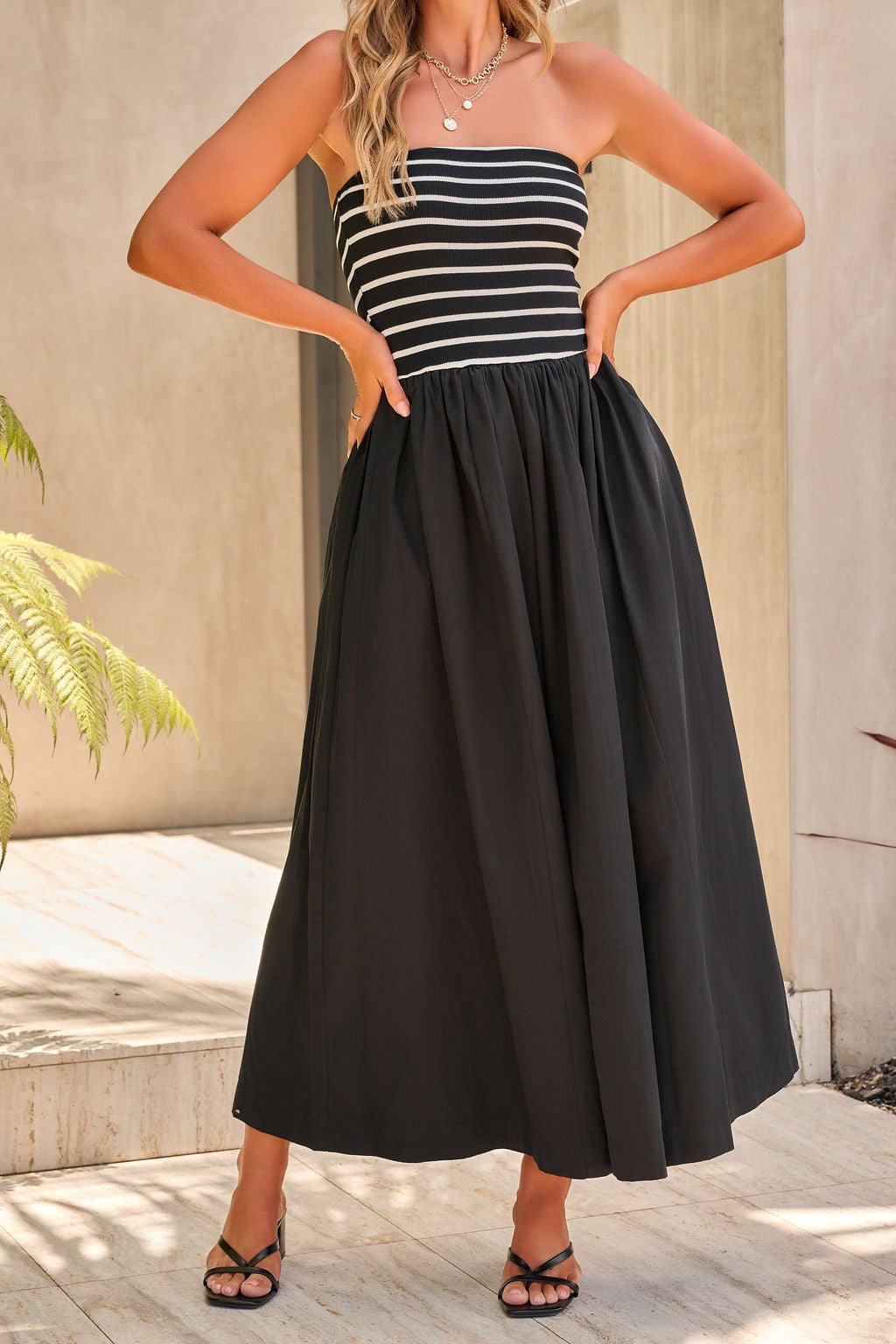 Woman in the Monaco Strapless Midi Dress - Black, hands on hips, standing on a sunlit patio. FINAL SALE.