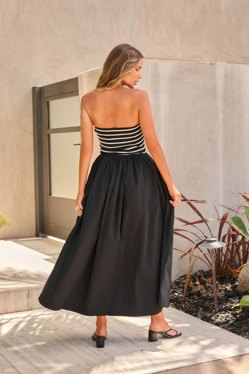 Woman in the Monaco Strapless Midi Dress - Black stands outdoors, facing away, holding her skirt.
