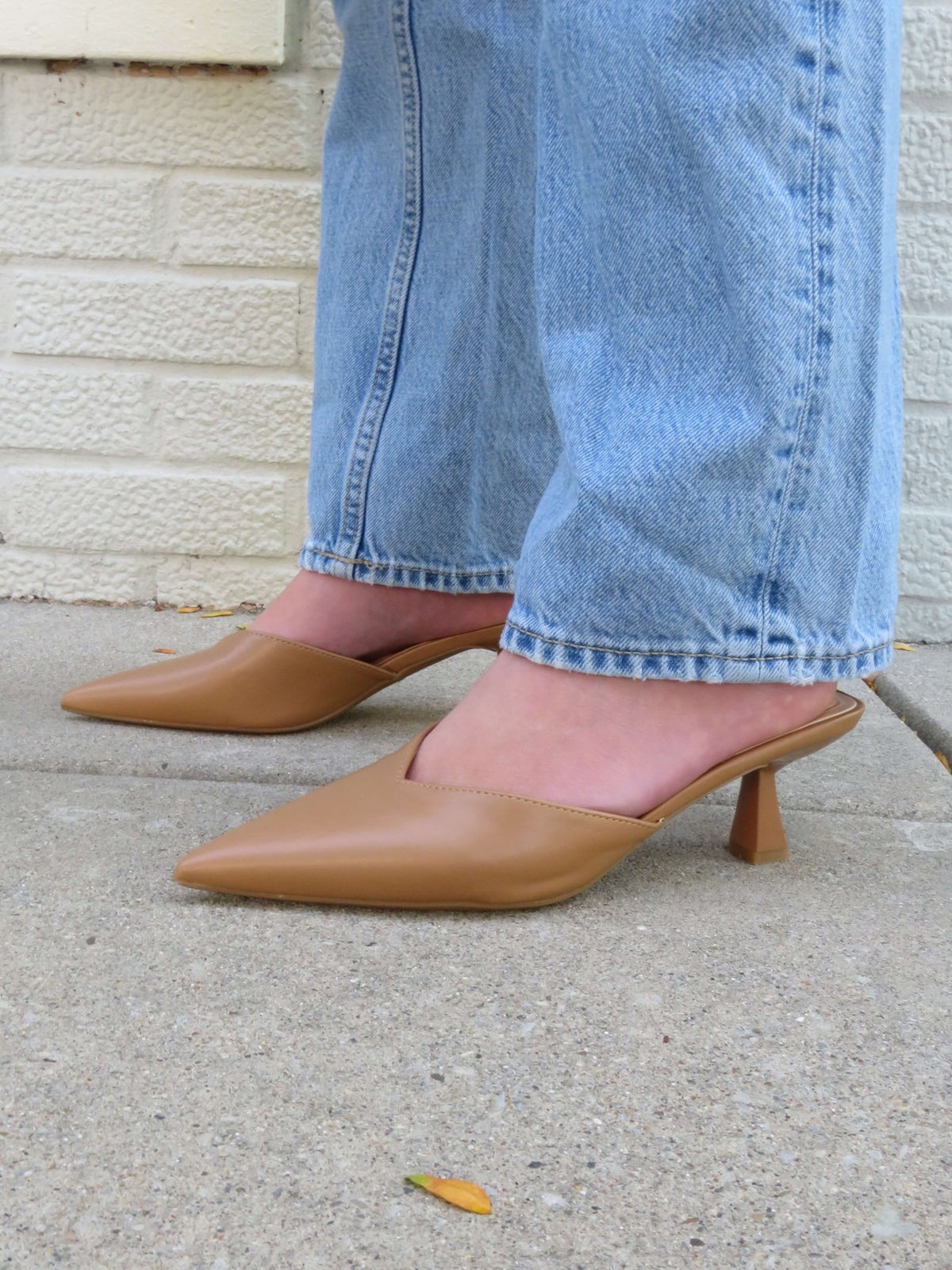 Person wearing Morley Camel Open Back Kitten Heels - FINAL SALE and light blue jeans on a concrete sidewalk near a cream brick wall.