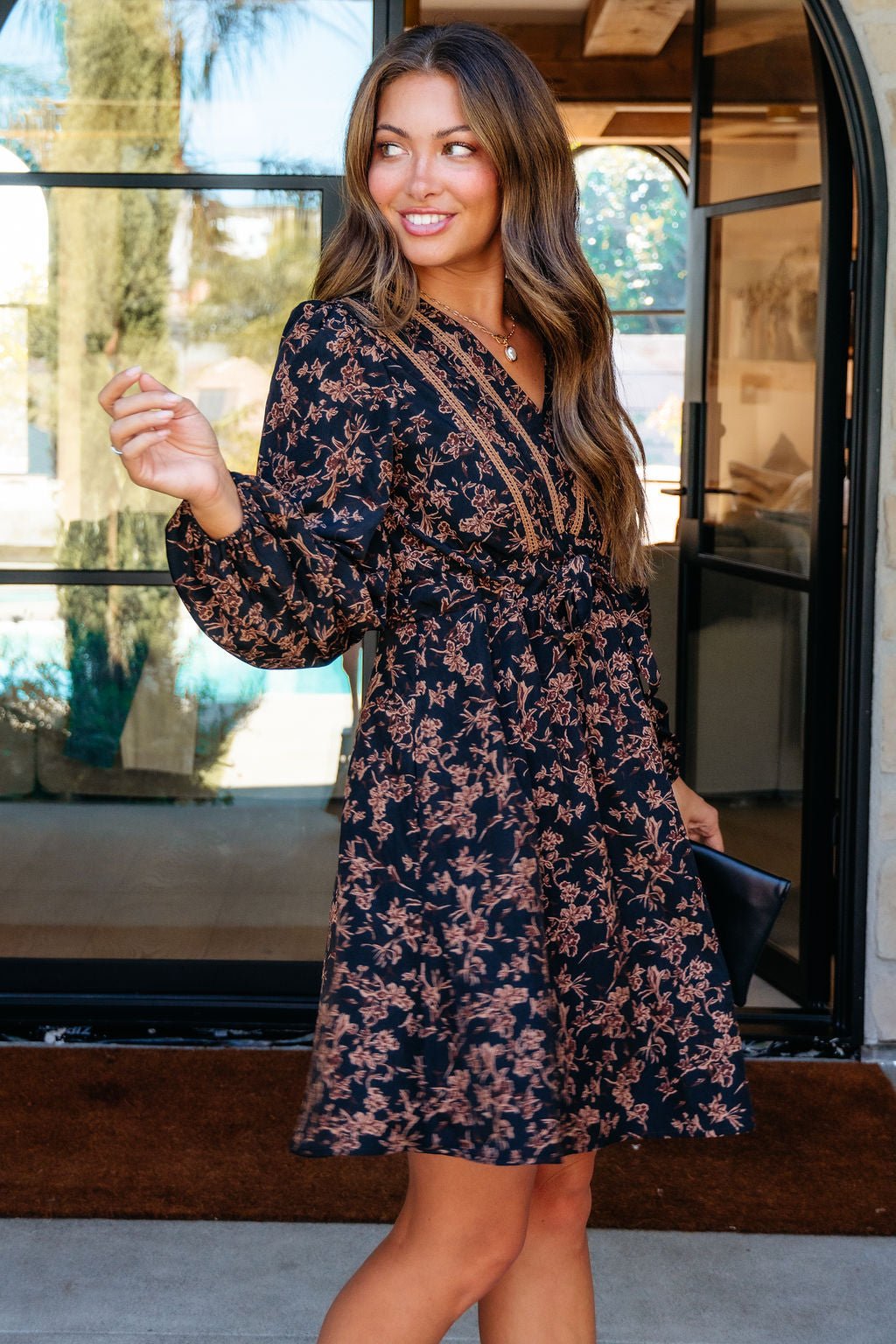 Woman in a Multi Black Floral Print Mini Dress stands by a glass door, smiling and holding a black clutch.