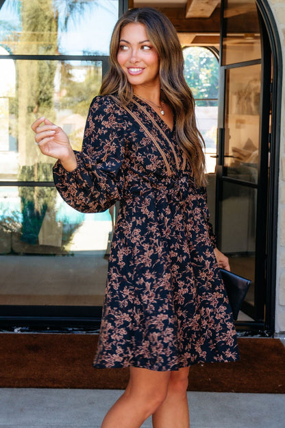 Woman in a Multi Black Floral Print Mini Dress stands by a glass door, smiling and holding a black clutch.
