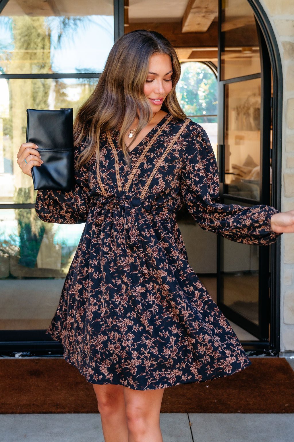 Woman models the Multi Black Floral Print Mini Dress and a black clutch outdoors by large glass doors.