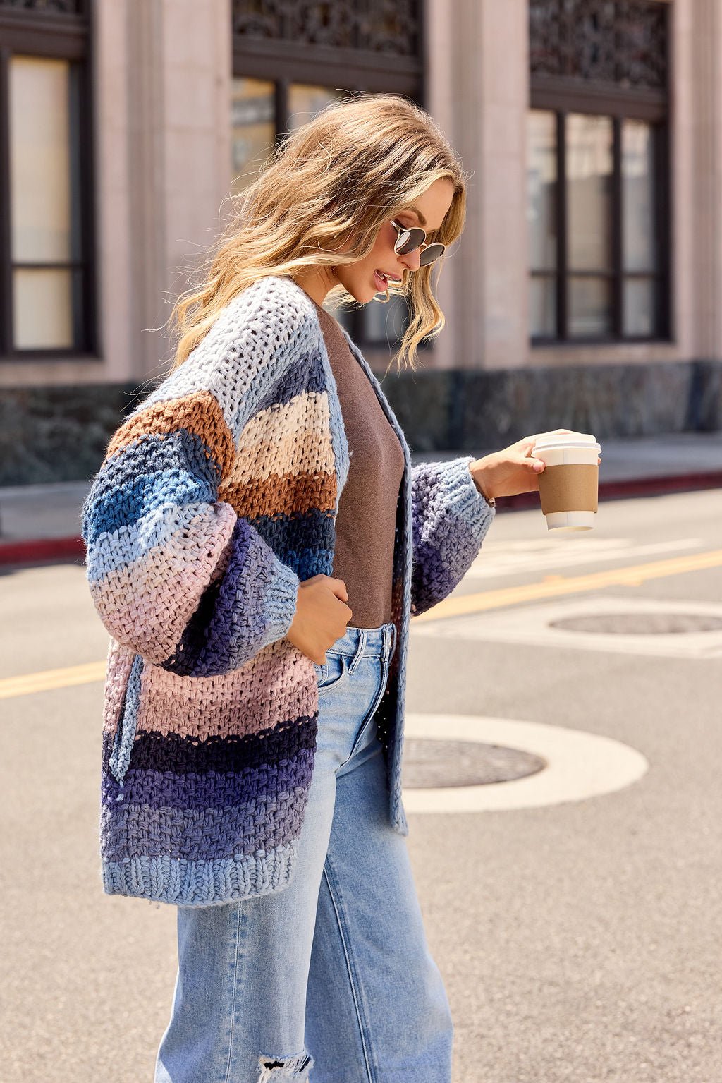 Woman in sunglasses wears a Multi Blue Stripe Crochet Cardigan and jeans, holding coffee while walking on a city street.
