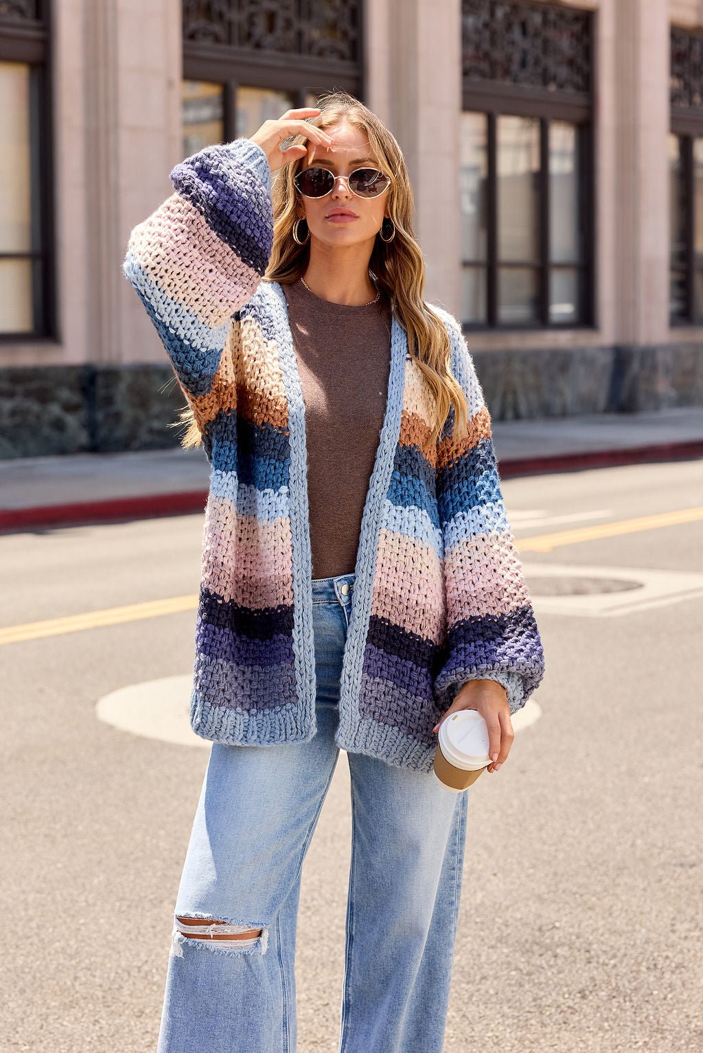 Woman in sunglasses wears a Multi Blue Stripe Crochet Cardigan, brown shirt, and ripped jeans while holding coffee on a city street.