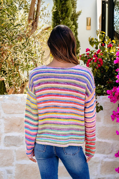 A woman with long hair wears the Multi Rainbow Striped Sweater with blue jeans, standing outdoors by a stone wall and flowers.