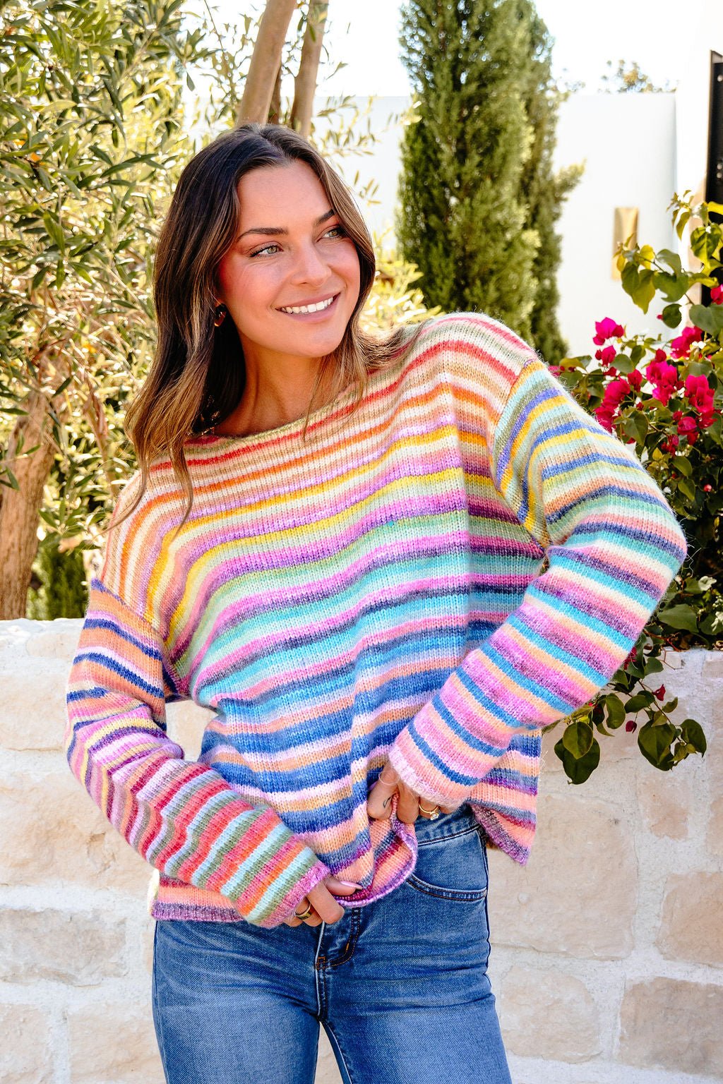 A woman in a Multi Rainbow Striped Sweater and jeans stands outdoors by greenery and a stone wall.