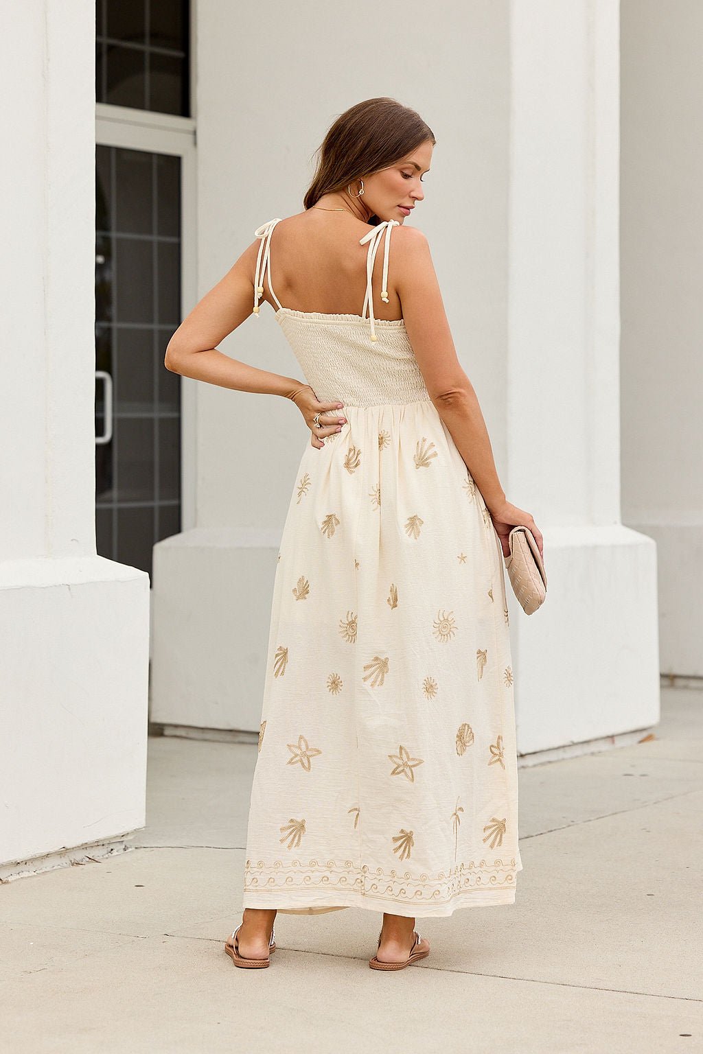 Woman in the Natural Beauty Embroidered Maxi Dress, holding a clutch and standing outdoors, faces away.