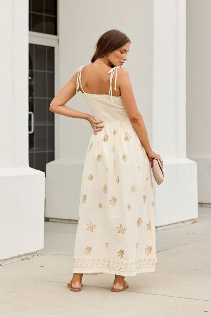 Woman in the Natural Beauty Embroidered Maxi Dress, holding a clutch and standing outdoors, faces away.
