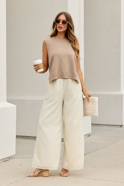 Woman in sunglasses wears Nine To Five Cashmere Sweater - Dark Beige with white wide-leg pants, nude heels, drink, and woven clutch.