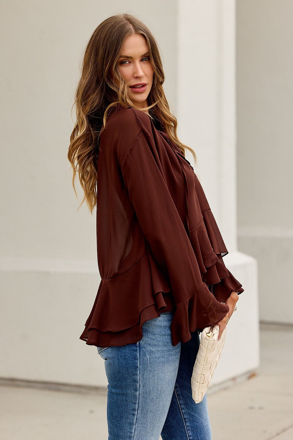 Woman with wavy hair in the Office Bell Sleeve Top - Brown FINAL SALE holds a white clutch, showing work-to-weekend style outside.