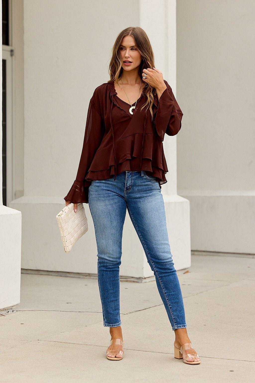 Woman in Office Bell Sleeve Top - Brown, blue jeans, and sandals carries a white clutch—work-to-weekend style on the sidewalk.