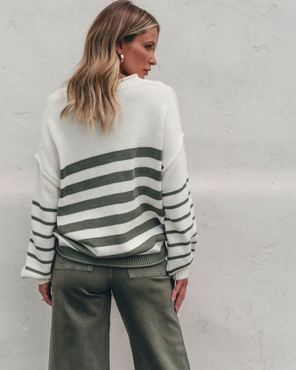 Woman in olive and cream striped pullover sweater with green pants stands against white wall, back to camera.