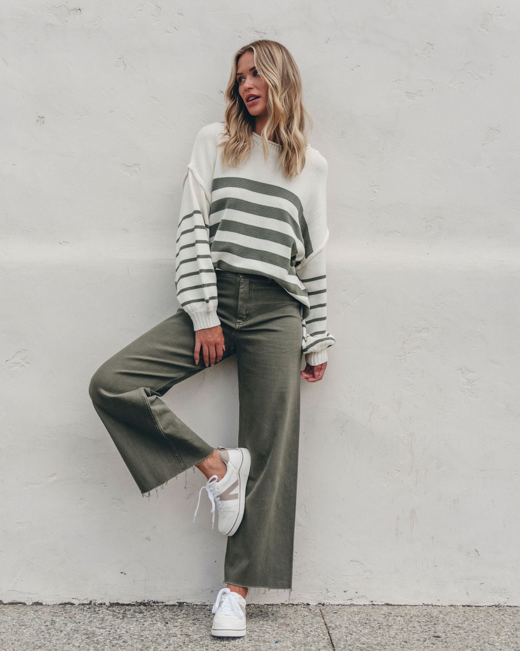 Woman wearing the Olive and Cream Striped Pullover Sweater leans against a white wall, showcasing a chic fall look.