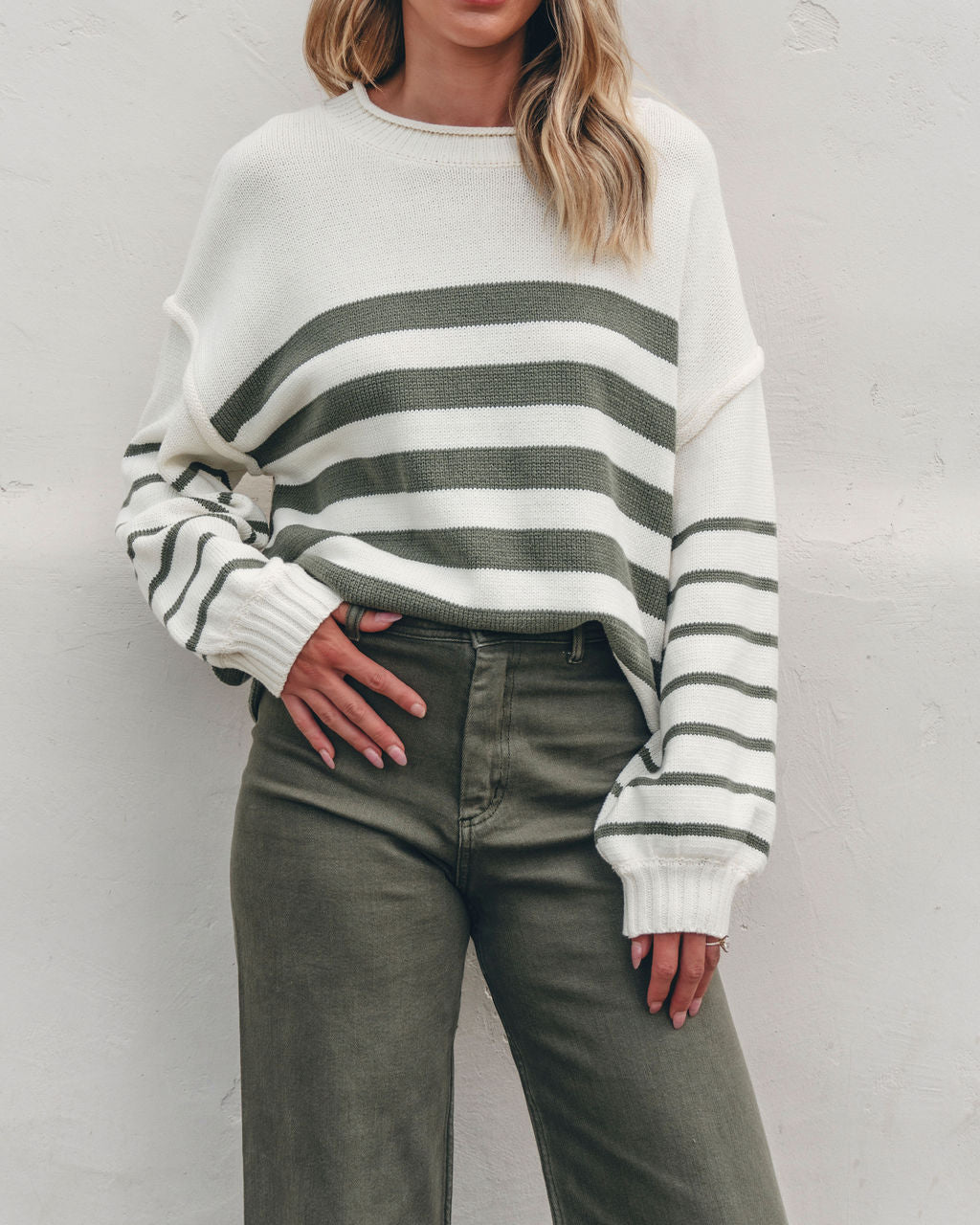 Model in Olive and Cream Striped Pullover Sweater with olive pants, a cozy fall look, stands against a white wall.