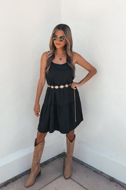 Woman in the On Point Tiered Mini Dress - Black stands with hand on hip, accessorized with tan boots, sunglasses, and a gold belt.